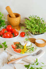 Poached egg with leaking yolk on zucchini and greens pancakes on a plate with pea microgreens and tomatoes with a cutlery on a serving table.