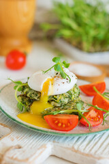 Poached egg opens with yolk leaking on zucchini and greens fritters on a plate of pea microgreens and tomatoes - close-up. Blurred background and foreground