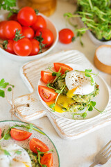 Poached egg with leaking yolk on zucchini and greens pancakes on a plate with pea microgreens and tomatoes on a served table.