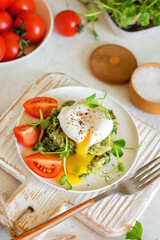 Poached egg with leaking yolk on zucchini and greens pancakes on a plate with pea microgreens and tomatoes on a served table.