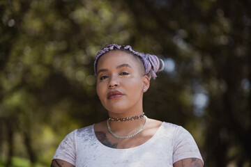 Posing in a leafy forest, a nonbinary model of multiracial African American and Latinx background exudes confidence. Their vibrant purple braids signify their queer identity and gender journey.