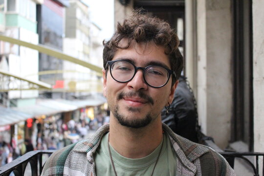 closeup portrait of young man with beard and glasses smiling with crowded city street view *4
