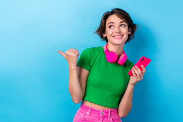 Photo of dreamy funky woman wear green t-shirt listening songs modern device pointing thumb empty space isolated blue color background