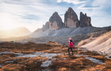 Walking girl with backpack on the trail in mountains at sunset in autumn. Tre Cime, Dolomites, Italy. Beautiful landscape with young woman, high rocks, path, stones, orange grass, sky in fall. Hiking - obrazy, fototapety, plakaty