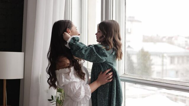 Beautiful young mom and adorable daughter embrace and enjoy spending time together by the panorama window at home. Happy motherhood, life value, affection, trust and parent love concept. Family day