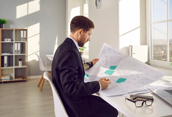 Side view portrait of a professional cartographer working with printed cadastral map at table on his workplace. Young man analyzing cadastral map and searching for a building plot. - obrazy, fototapety, plakaty