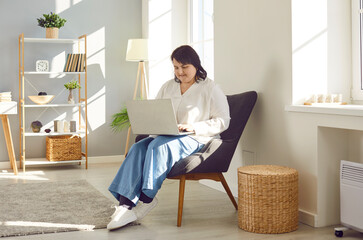 Portrait of a plus size young smiling girl sitting in the living room at home using laptop for...