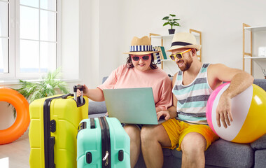 Happy family couple in summer hats and sunglasses sitting on sofa with beach ball and travel bags, using laptop computer, and booking tickets and holiday accommodation online - obrazy, fototapety, plakaty
