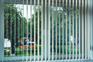 Venetian blind hanging on large window of house with garden view. Rolling shutter is opening.
