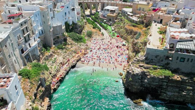 Lama Monachile Cove Beach with clear turquoise water by dramatic rugged cliffs. Aerial drone view of Polignano a Mare with tourists relaxing enjoying and having good time in summer by the beach, Italy