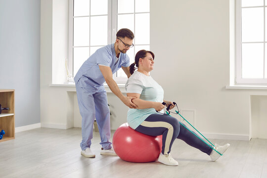 Nurse Man Helping Fat Patient To Do Sport Stretching Fitness Exercises With Rubber Band Sitting On Fit Ball In Clinic. Male Physiotherapist Or Health Care Worker Helping Overweight Woman In Rehab.