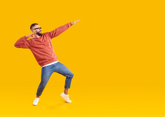 Fototapeta na wymiar Cheerful man having fun showing dance moves in dab style on orange background in studio. Caucasian man in youth casual clothes jokingly fools around near copy space. Fun concept. Full length.