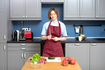 Young attractive housewife in red apron slicing vegetables and preparing vegetarian food