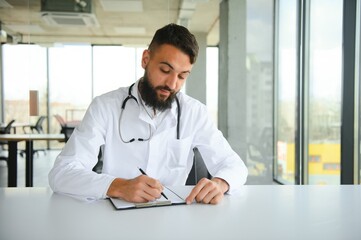 Portrait of arabic doctor handsome young man in workwear posing at modern clinic, sitting at workdesk