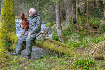 The hiker and Irish Setter sit together on a log by the riverbank. The peaceful scenery and the...