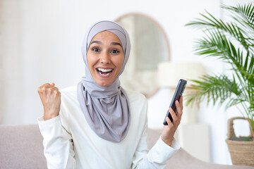 Muslim young woman in hijab looking surprised at camera and happy, holding phone in hands, got good news. messages, financial success.