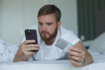 Young happy man doing online shopping with smartphone, lay in bed in bedroom at home, hold in hand credit bank card and cell mobile phone and smile