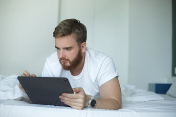 Young happy man use tablet laptop computer at home in bed, lay in bedroom, looking at screen of gadget. Concept of freelance work, distant job, online shopping or education