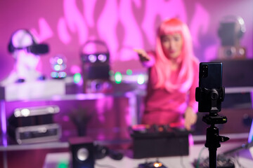 Asian dj with pink hair performing techno music using professional mixer console while recording process with mobile phone camera. Smiling artist playing song during night time in club