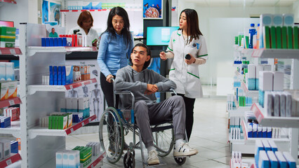 Young man wheelchair user talking to pharmacist about medicaments, asking for prescription treatment or medicine. Person with disability and caretaker receiving supplements and vitamins in pharmacy.