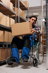 Asian man wheelchair user carrying parcel in postal warehouse aisle full of cardboard boxes on...