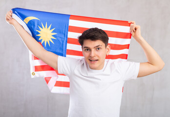 Happy young man with flag of Malaysia in hands posing against light unicolored background