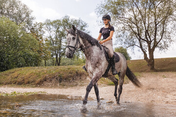 Girl riding gray horse down the calm river water with forest greenery reflections from the...