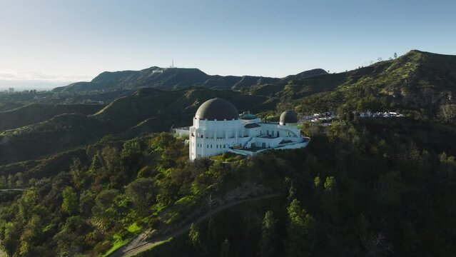 Los Angeles city California USA March 2023. Flying around dome architecture Griffith Observatory, Hollywood sign on bright green hill at sunset. Aerial scenic landmark buildings in green spring park