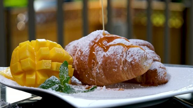 watering croissant with liquid caramel on white plate mango mint leaves and delicious french dessert breakfast lunch thin stream of sweet syrup pouring on bun pastries fresh food on hotel balcony