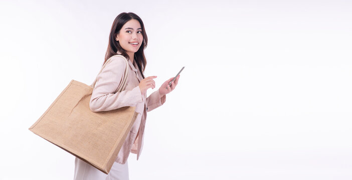 Cheerful shopper woman carry eco tote handbag holding mobile phone looking copy space standing on isolated white background. Happy asian mixed race girl holding shopping fabric bag pointing smartphone