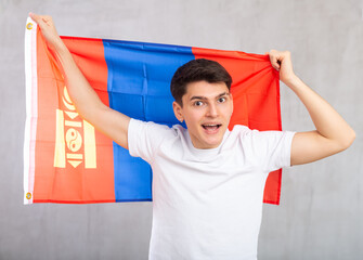 Enthusiastic young man holding large flag of Mongolia against light shadeless background