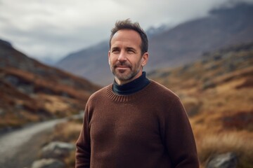 Photography in the style of pensive portraiture of a grinning man in his 40s wearing a cozy sweater against a mountain landscape background. Generative AI