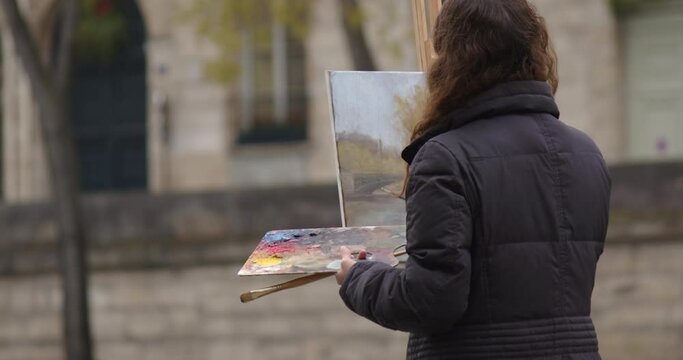 Woman painting a picture near the river Seine. Creative woman artist paints a picture with a brush while standing near the easel in Paris. Woman artist, art for sales, inspiration