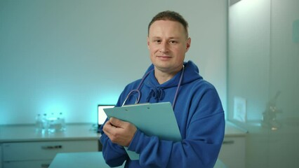 Medium shot of young smiling male vet with folder in hands and stethoscope around neck standing in laboratory in modern vet clinic. Man looks into camera. High quality 4k footage