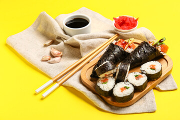Fototapeta na wymiar Wooden board with tasty sushi cones, rolls, soy sauce, ginger and garlic on yellow background