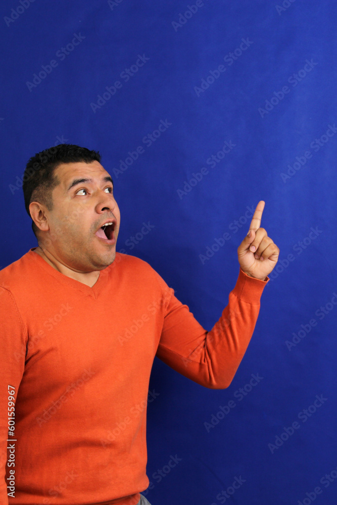 Wall mural Adult man forty years old latino brunette with orange sweater points with his hands blue background copy space with happy and natural expression