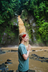The girl is meditating, her hands are folded in namaste, in the jungle against the backdrop of a waterfall.