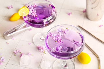 Glasses of cocktail with lilac flowers and kumquat on light tile table, closeup