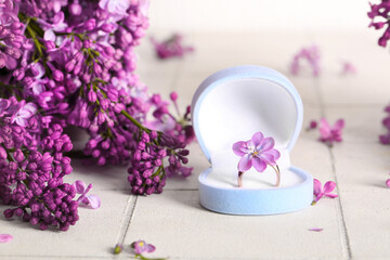 Fototapeta na wymiar Box with engagement ring and beautiful lilac flowers on light tile table