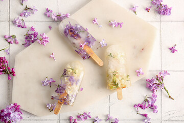 Board with beautiful floral popsicles and lilac flowers on light tile background