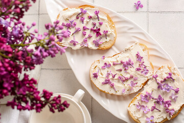 Fototapeta na wymiar Plate of sandwiches with tasty cream cheese and lilac flowers on light tile table, closeup