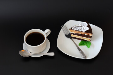 A cup of hot black coffee on a saucer with a spoon and a plate with a piece of biscuit puff chocolate cake with mint on a black background.