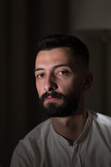 Dramatic lighting illuminates a handsome young Syrian man sitting indoors. His facial hair, dark eyes, and serious expression create an air of mystery.