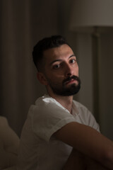 Sitting in a reading chair next to drawn curtains and a floor lamp, a young man from Syria looks over to gaze directly at the camera. His dark eyes and thick beard adding drama to his handsome face.