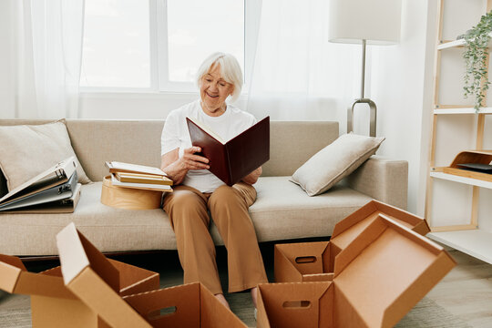 elderly woman sits on a sofa at home with boxes. collecting things with memories albums with photos and photo frames moving to a new place cleaning things and a happy smile.