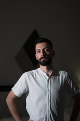 Naklejka premium With a serious expression, a young Syrian man dressed in a white polo shirt poses in a dimly lit room. His black beard and handsome features are illuminated by a single light source.