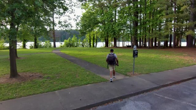footage of an African American man wearing a backpack, holding a camera hiking in a park with lush green trees and grass at sunset at Proctor Landing Park at lake Acworth in Acworth Georgia USA