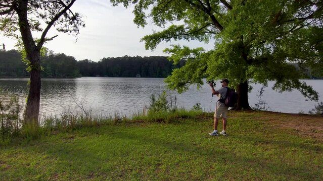 footage of an African American man wearing a backpack, filming on the banks of a lake with lush green trees and grass at sunset at Proctor Landing Park at lake Acworth in Acworth Georgia USA