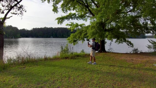 footage of an African American man wearing a backpack, filming on the banks of a lake with lush green trees and grass at sunset at Proctor Landing Park at lake Acworth in Acworth Georgia USA