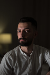 Wearing a white polo shirt, a young Syria man in a dimly lit room poses for a portrait. He has a somber expression, handsome features, and a black beard.